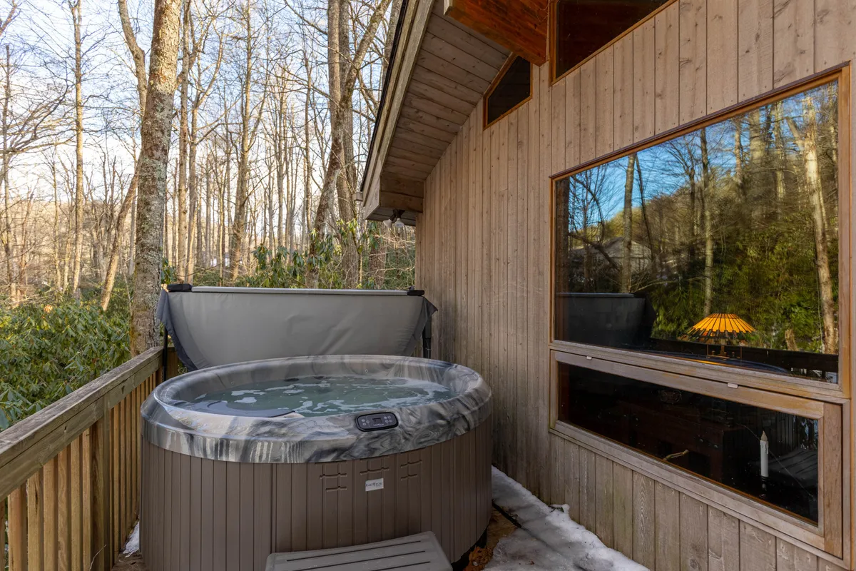 This exterior image features a bubbling hot tub on a wooden deck framed by surrounding trees and a large window, creating a sense of privacy and immersion in nature. The tranquil setting encourages relaxation after outdoor adventures, with space for group soaking and nature views.