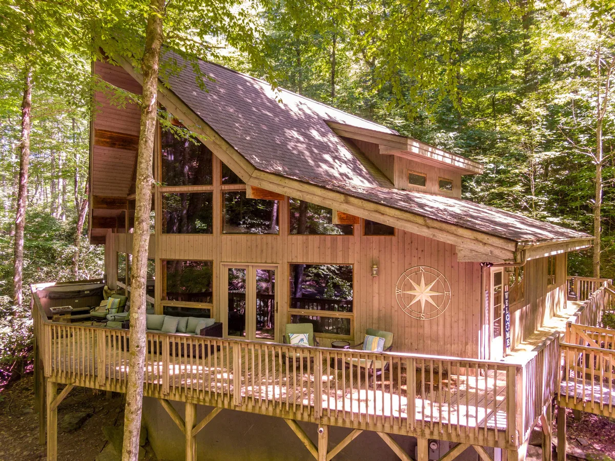 The image showcases the chalet-style home's striking wooden architecture, featuring expansive 20-foot-high windows that flood the interior with light. The spacious wooden deck is inviting, accented by a compass rose decoration, and surrounded by serene woods that hint at the privacy and natural beauty of the setting. The lush forest backdrop and glimpses of the creek create an atmosphere of peaceful seclusion, perfect for relaxing or gathering with loved ones.