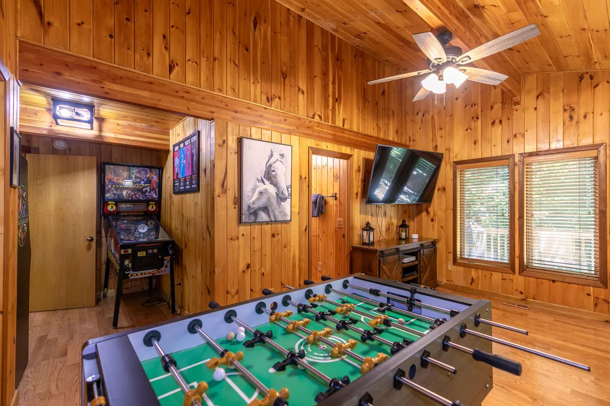 This image showcases a vibrant game room featuring a foosball table, an arcade machine, a TV, wooden paneling, and playful artwork. The space is set up for multiple gaming activities and has an inviting, fun-filled ambiance.