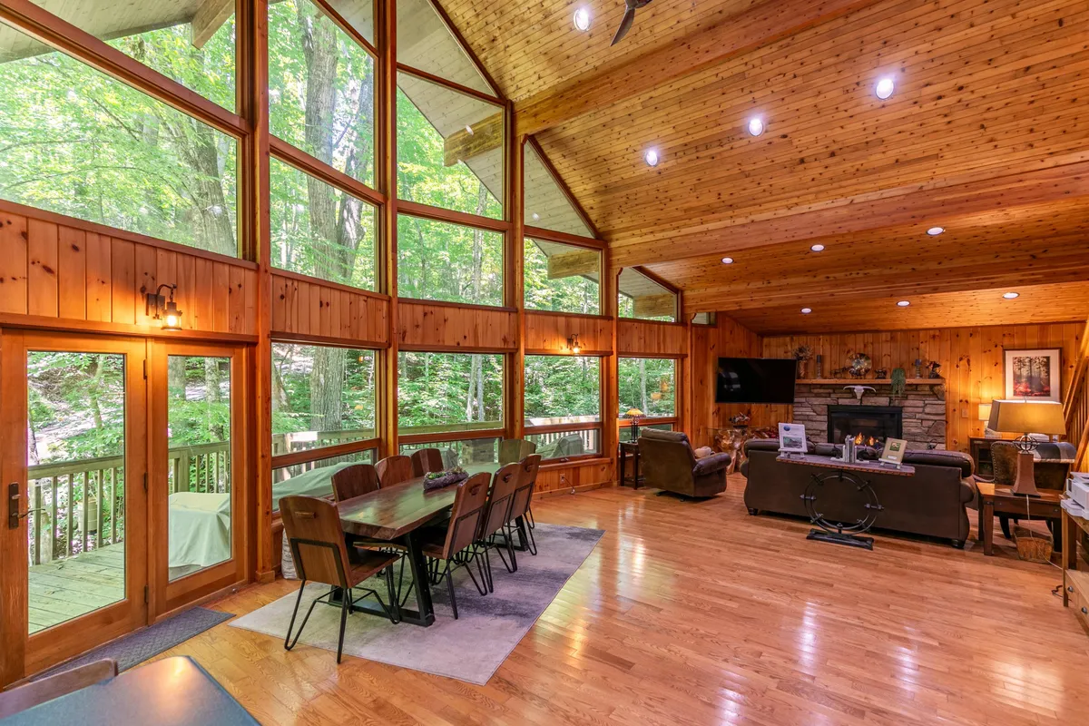 A welcoming open-concept living area featuring large windows with a sweeping forest view, a striking stone fireplace, a spacious dining table, and a comfortable seating area. The room is bathed in natural light, showcasing the seamless flow from living to dining space, perfect for gatherings or relaxing evenings.