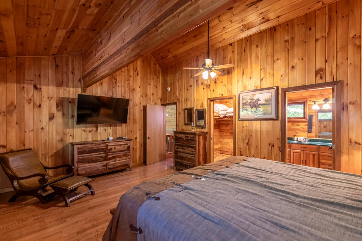 A warmly lit king bedroom featuring wooden walls, a plush bed with textured bedding, mounted TV, rustic furniture, and decorative artwork. The space feels cozy and private, ideal for relaxation and restful nights.