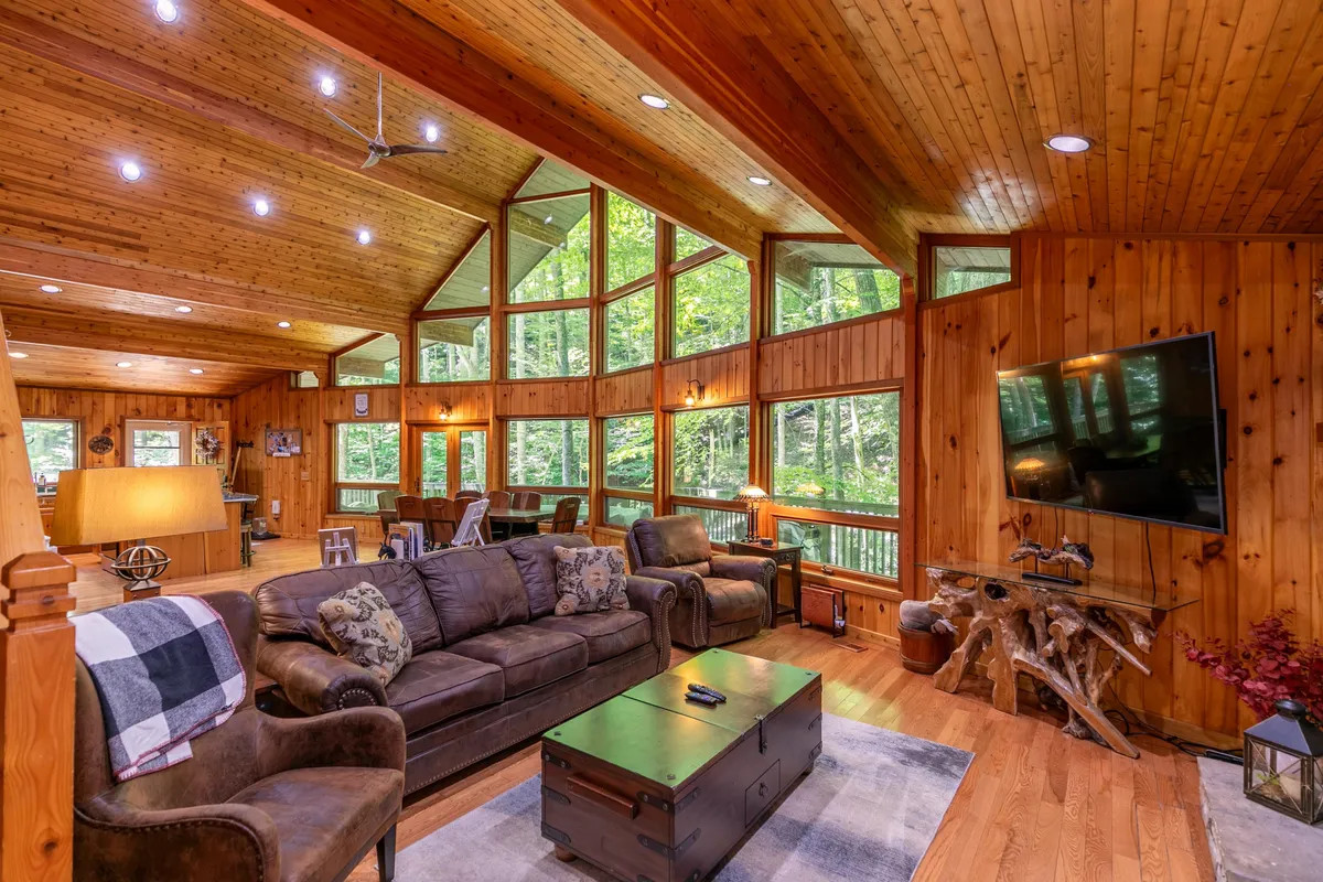 A cozy living room anchored by a comfortable sofa, rustic wooden ceiling, large windows framing forest views, a decorative table, and a sizable TV. The inviting setup encourages family movie nights or serene mornings spent gazing outdoors.