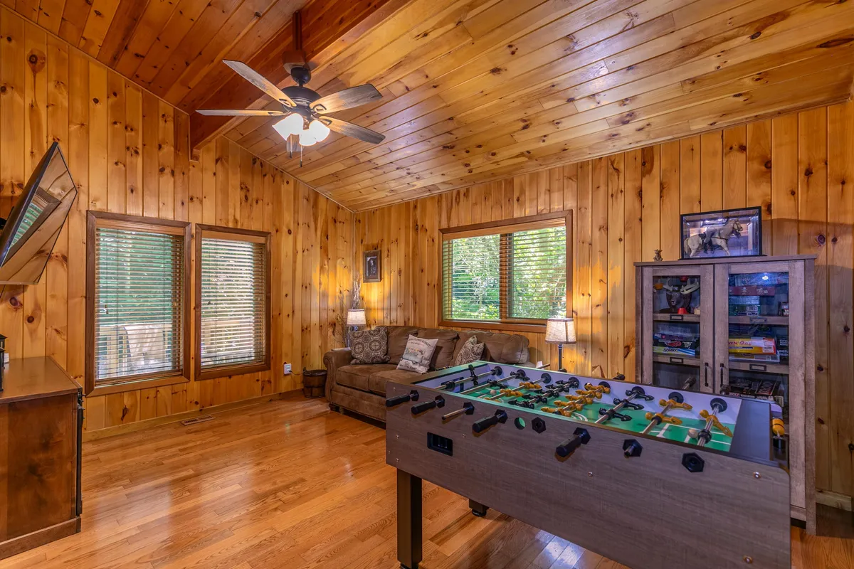 A cozy game room with a foosball table, inviting sofa, wooden walls, and large windows allowing in natural light. The arrangement encourages both play and relaxation, perfect for group gatherings or family fun.