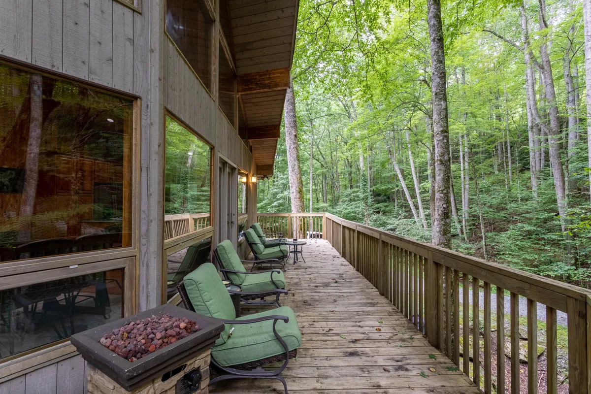 A wooden deck with green chairs and a fire pit is nestled among mature trees. The scene is serene, ideal for socializing, relaxing, or quietly appreciating the surrounding woods at sunset or sunrise.