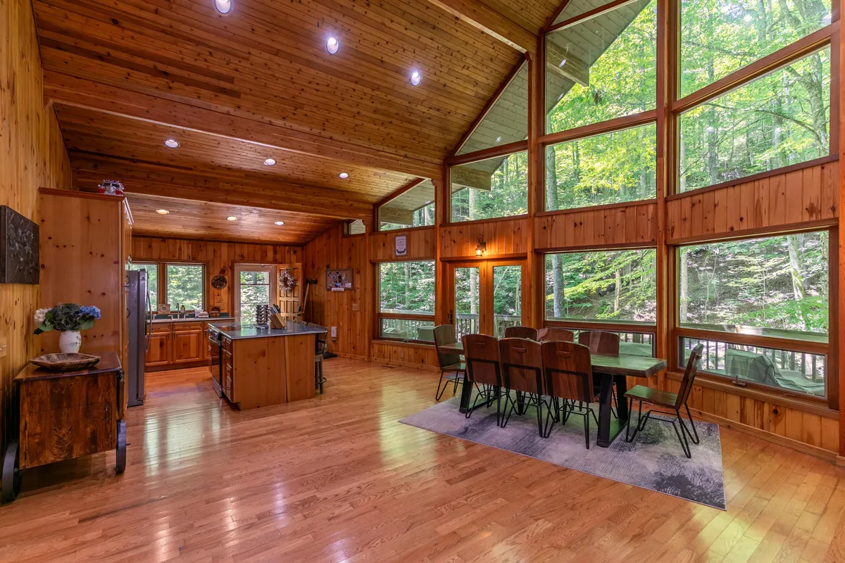 An inviting view of the dining area and modern kitchen, with a large kitchen island, expansive windows overlooking lush woods, and a dining table ready for shared meals. Sunlight fills the space, highlighting both the functional and social elements of the kitchen and dining zones.