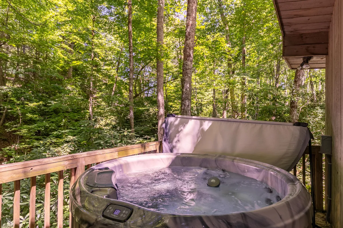 An inviting outdoor hot tub sits on a private deck surrounded by tall trees and wooden railings, offering a peaceful and secluded spot for relaxation. The view immerses guests in the natural beauty of the forest, perfect for unwinding under the stars.