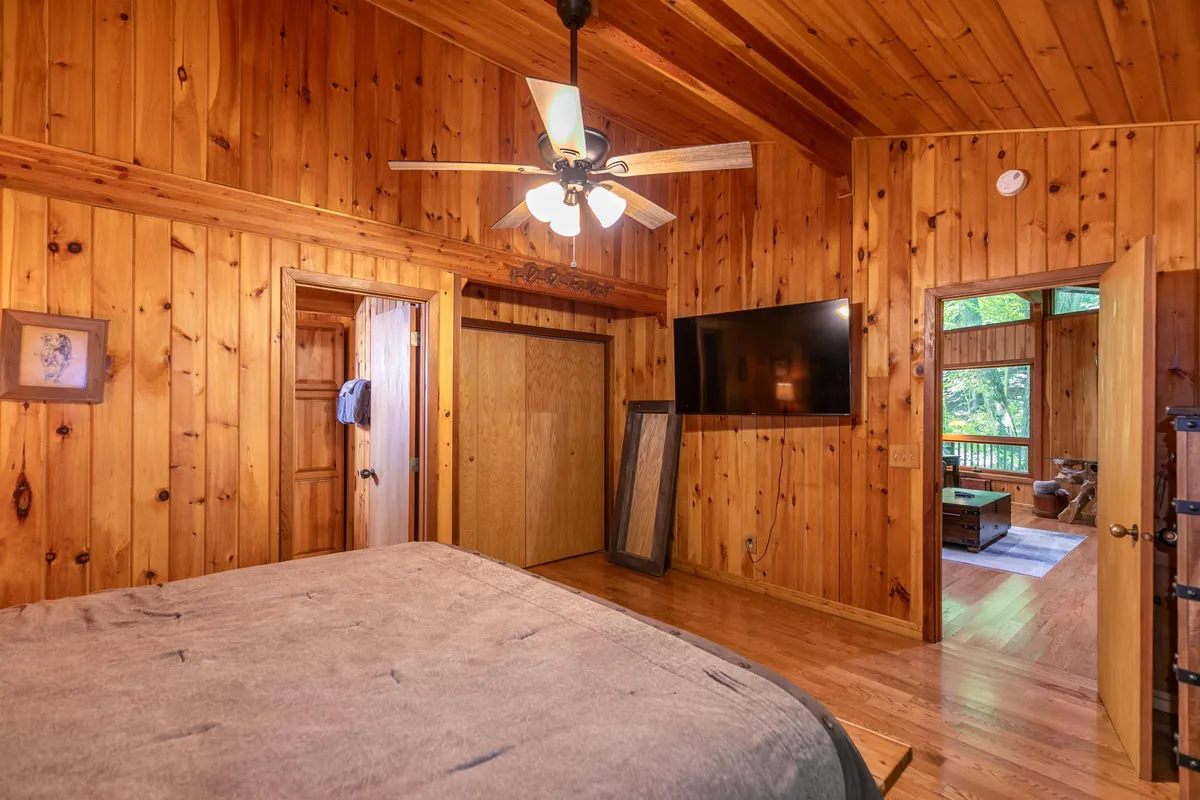 A king bedroom with rich wood-paneled walls, ceiling fan, large bed, and a TV mounted on the wall. The room is bathed in natural light from a window, suggesting views of the outdoors and a tranquil environment.