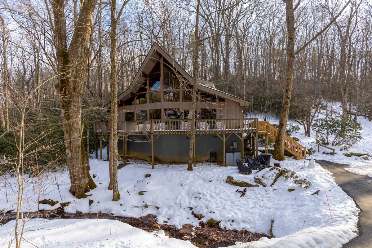 A wooden deck flanked by large windows faces a snowy, wooded landscape. The view captures the cabin’s cozy mountain setting during winter, suggesting magical, snow-draped mornings or evenings spent watching snowfall from the comfort of the retreat.