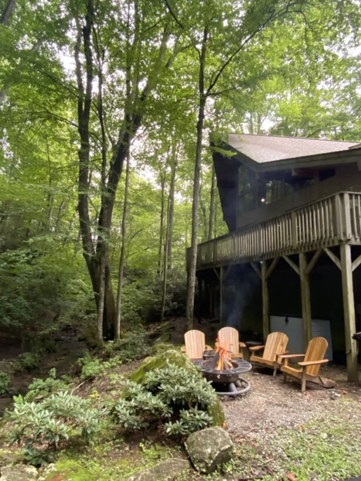 An inviting fire pit area is pictured under a leafy tree canopy, surrounded by Adirondack chairs with the upper deck of the house visible in the background. The warm glow of the fire pit sets a social atmosphere ideal for evening gatherings, storytelling, and stargazing with friends.