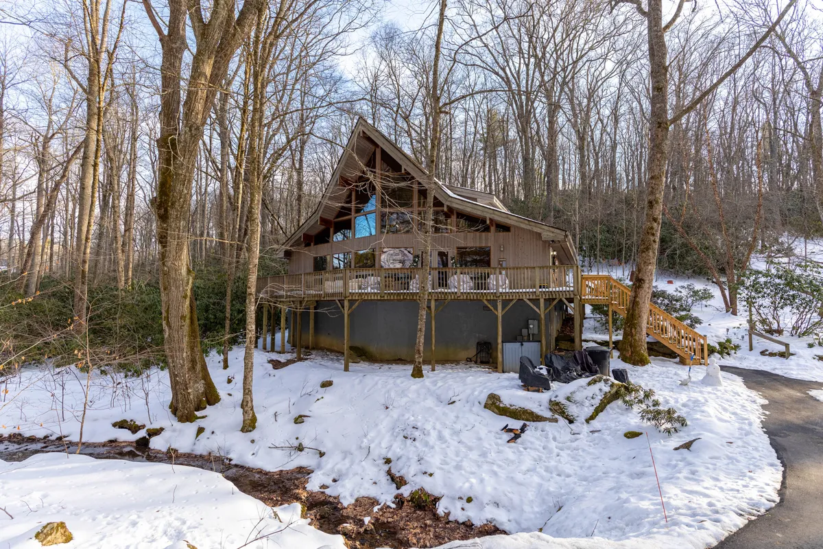 A broad exterior shot reveals the A-frame house nestled among snow-covered ground and mature trees, with a welcoming wooden deck. The image suggests ample privacy, space for outdoor play, and a tranquil retreat in a beautiful natural environment—ideal for group arrivals and shared adventure.