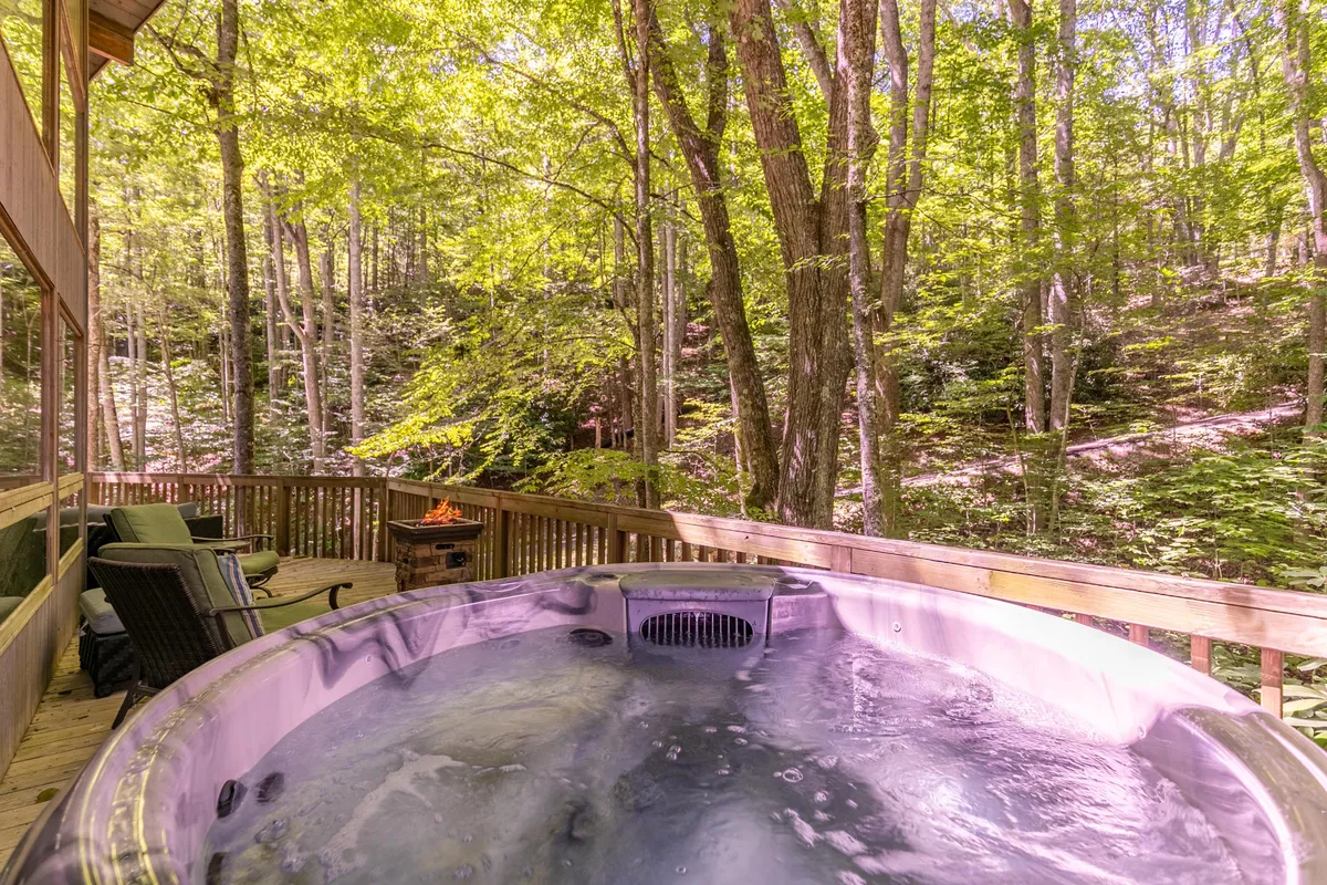 A tranquil outdoor scene featuring a wooden deck with a hot tub, surrounded by tall trees, and a nearby fire pit. The area is designed for relaxation and social gatherings, inviting guests to soak in the hot tub or gather around the fire under the stars.