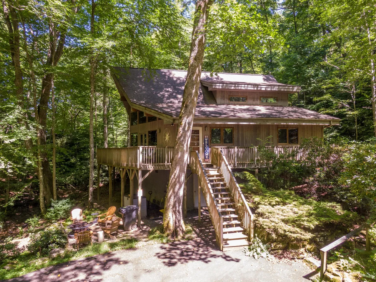 A spacious wooden deck overlooks a serene wooded area, featuring a staircase leading to a cozy fire pit surrounded by trees. The setting provides ample space for outdoor gatherings, enjoying peaceful coffee mornings, or romantic evenings by the fire.
